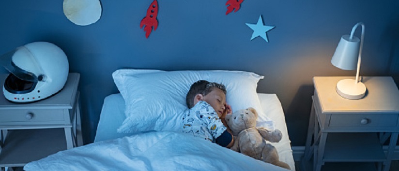 High angle view of little boy dreaming of becoming an astronaut while sleeping with teddy bear in space decorated room. Top view of dreamer child sleeping on bed with solar system and planet decoration during the night. Cute kid with astronaut helmet on side table and the light on.